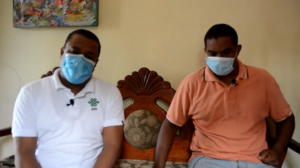 Two men sitting in front of the camera while wearing a facemask